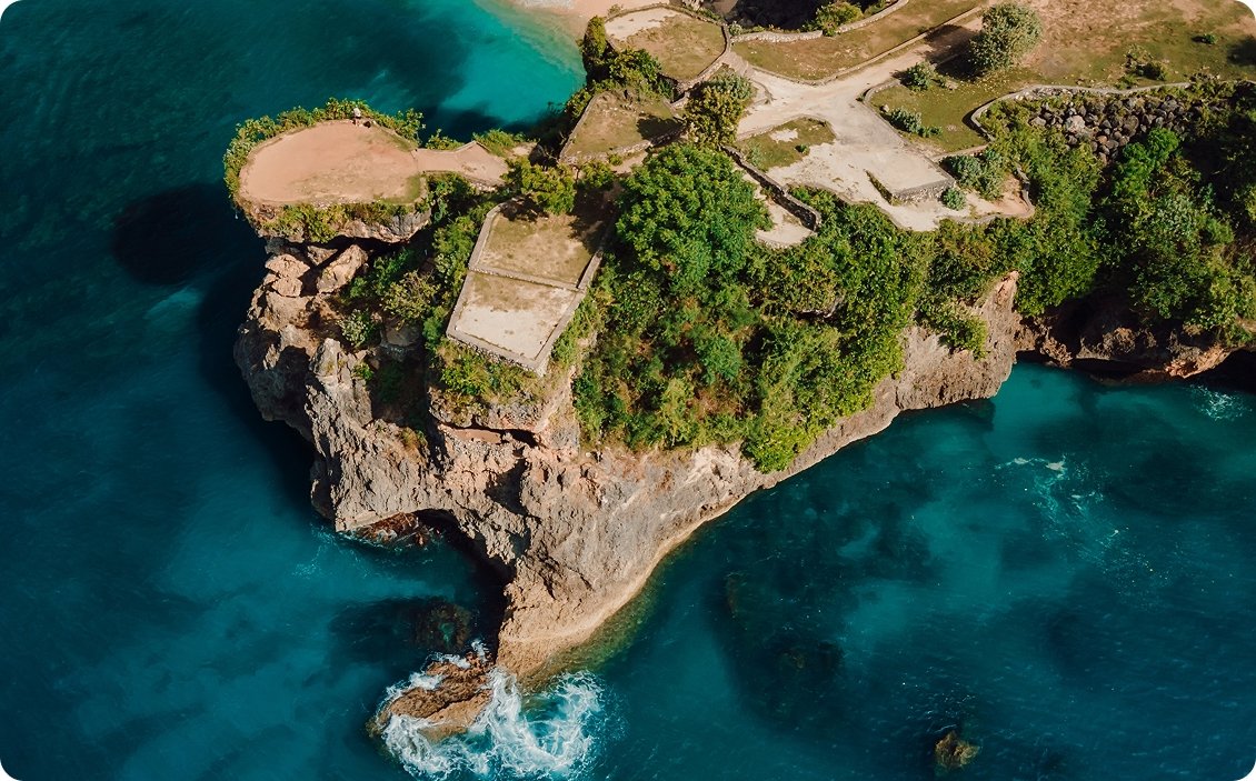 Top-down aerial view of a rugged rocky cliff headland in Bali, covered with lush green vegetation, jutting into the deep turquoise and blue waters of the ocean.
