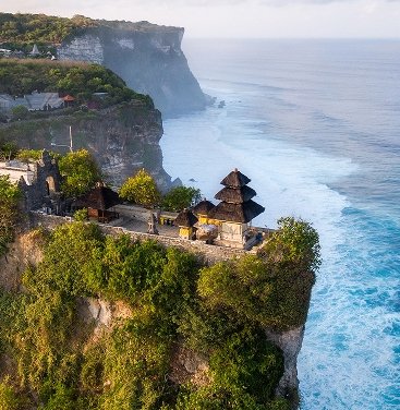 erial view of Pura Luhur Uluwatu temple perched on a dramatic cliff overlooking the Indian Ocean in Bali, with lush greenery and crashing waves.